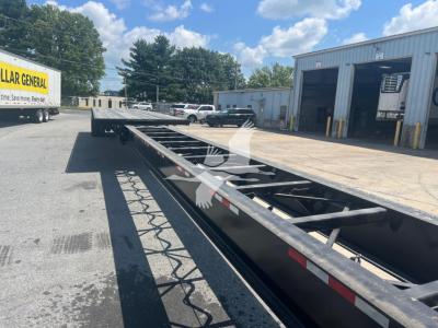 Extendable flatbed trailer by Manac with air ride suspension, long low-profile frame, visible stakes and frame lattice, parked outdoors with loading dock in background.