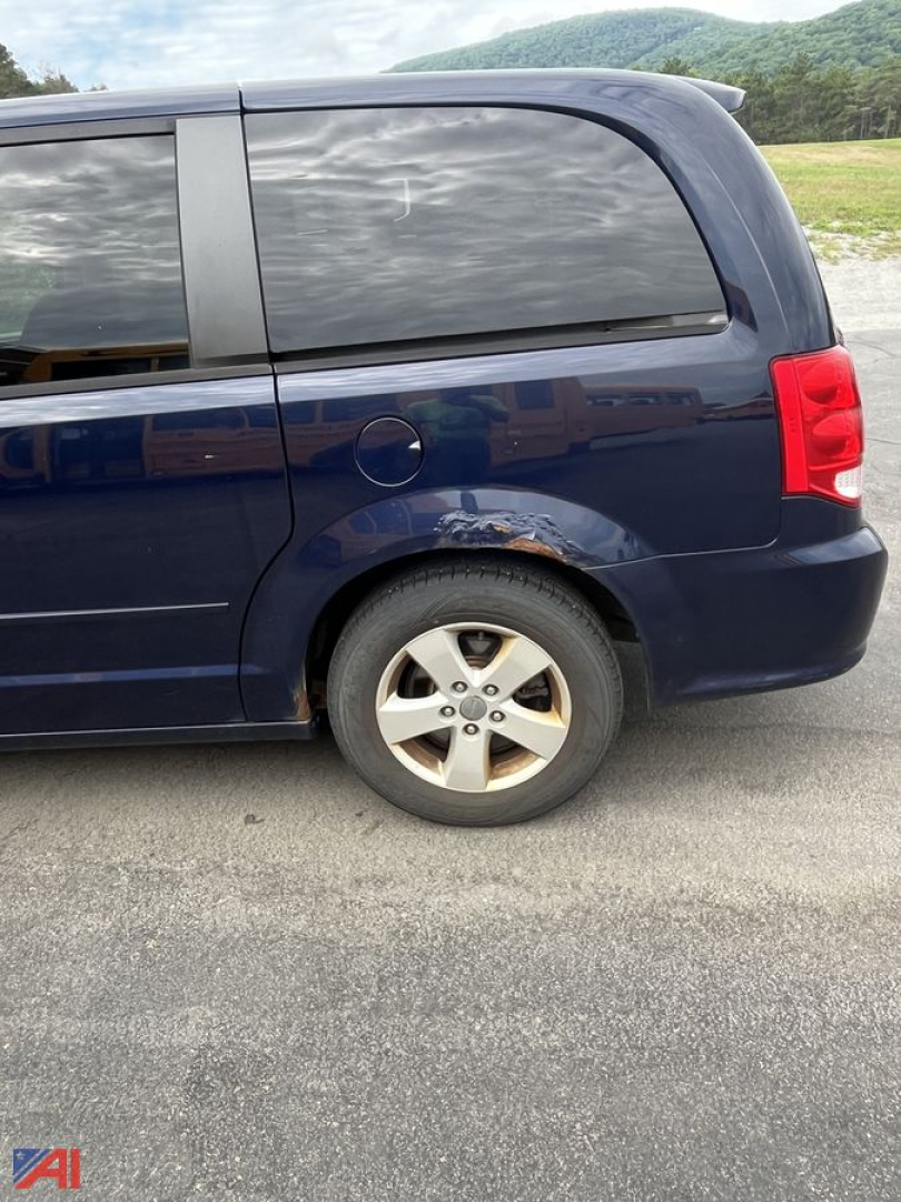 Vehicle side view showing rear quarter panel with wheel, blue Dodge Grand Caravan minivan, minor rust under rear door window.