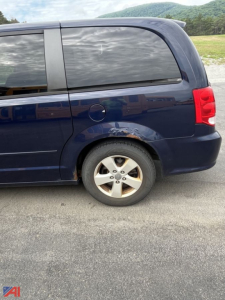 Vehicle side view showing rear quarter panel with wheel, blue Dodge Grand Caravan minivan, minor rust under rear door window.