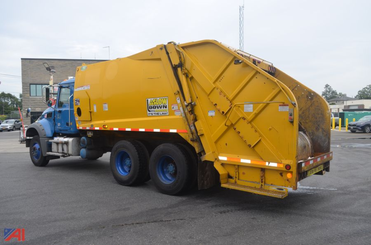 Rear/side view of a yellow garbage truck attached to a blue Mack truck cab. Side shows compacting body, hydraulic components, and warning decals.