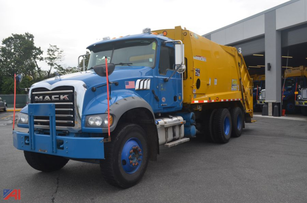 Mack Granite model truck in blue with yellow waste-hauler body, front view, VIN 1M2GR6GC3LM001675 visible, urban service yard.