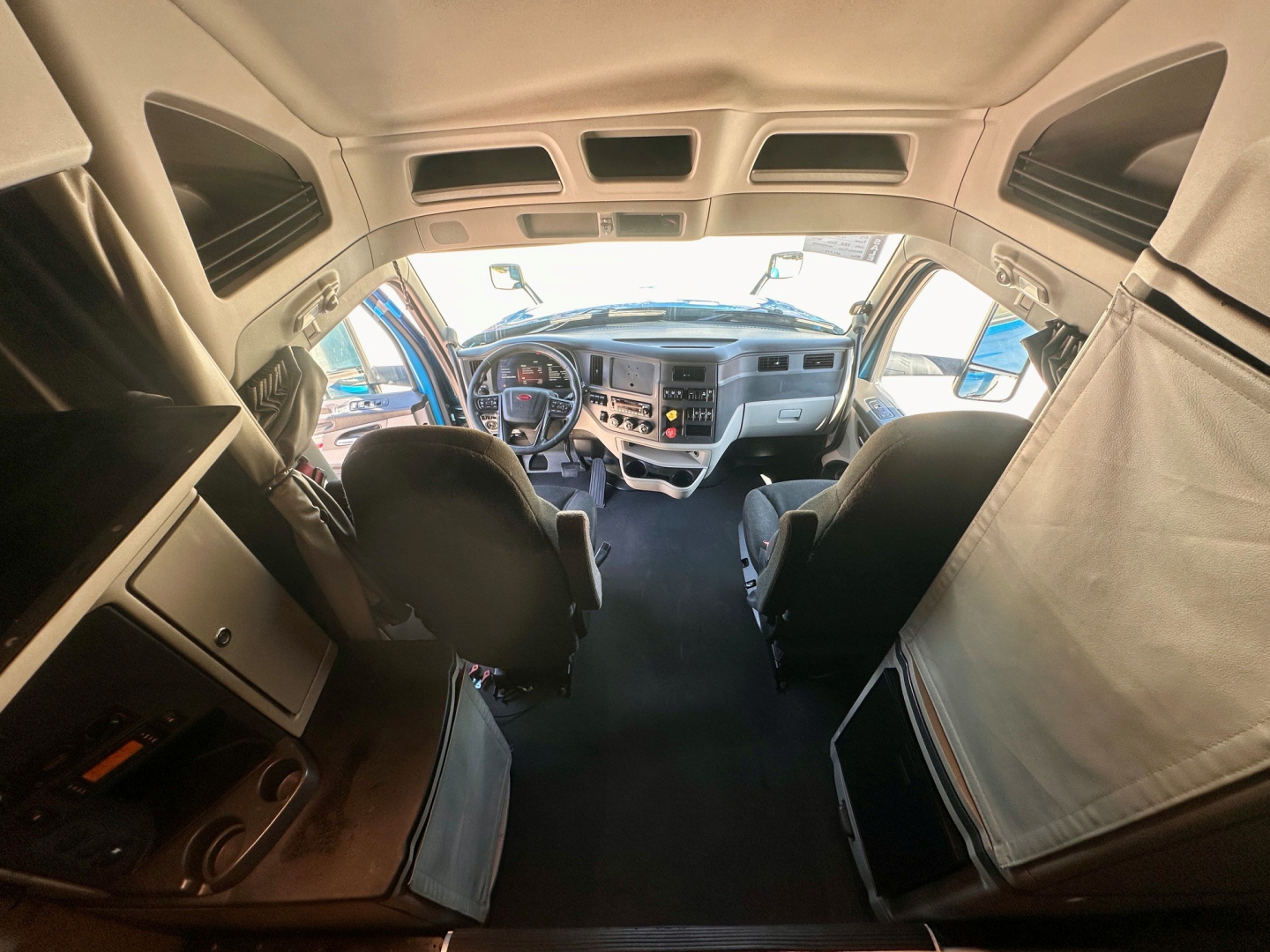 Truck cab interior of a 2022 Peterbilt 579, front windshield view from rear of sleeper with steering wheel center, dashboard gauges, two seats, overhead compartments.
