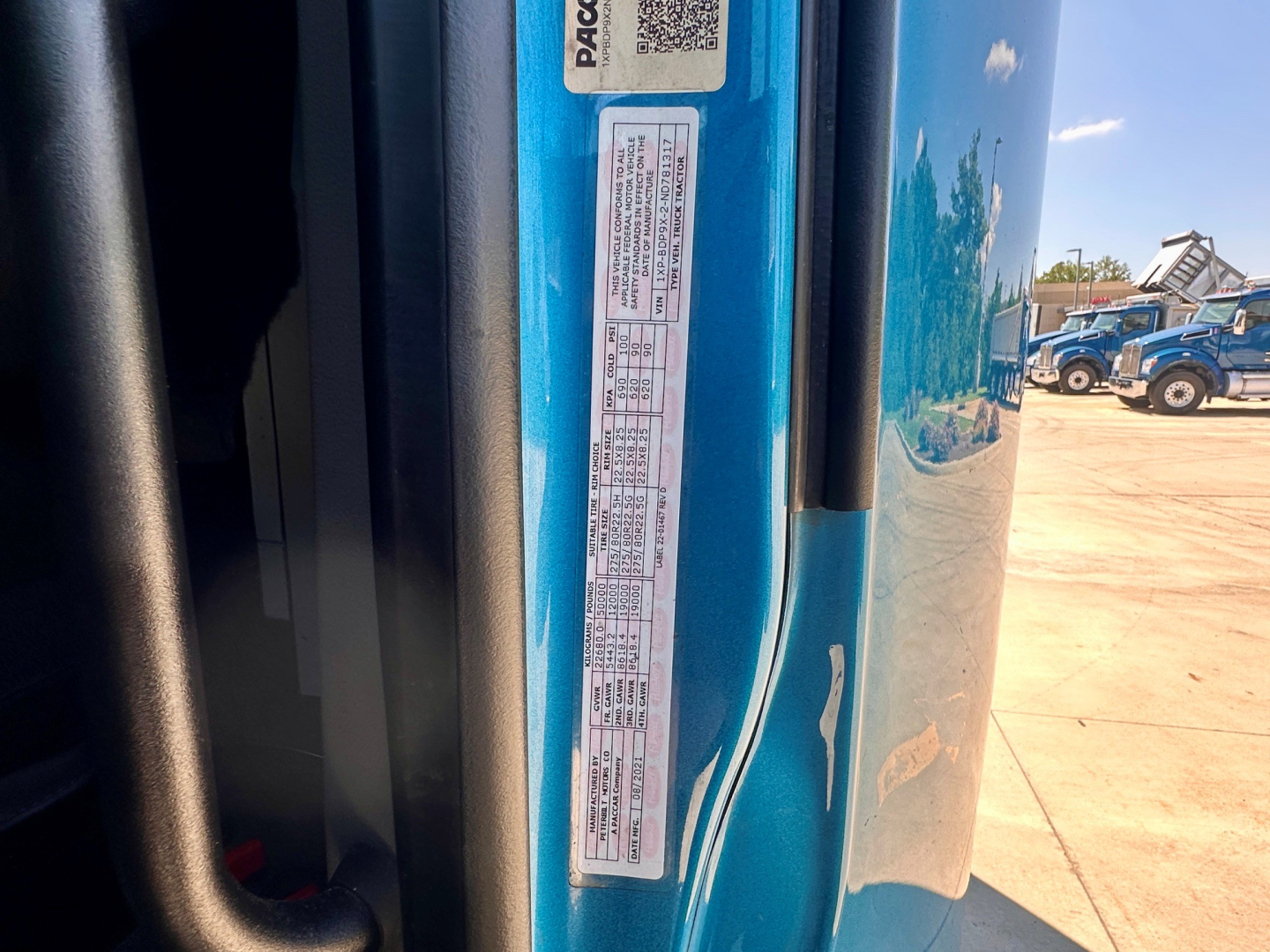 Close-up view of a blue Peterbilt truck door jamb with a vertical sticker containing vehicle information labels, text includes VIN barcodes and small print; portion of the cab and exterior reflect in the background.