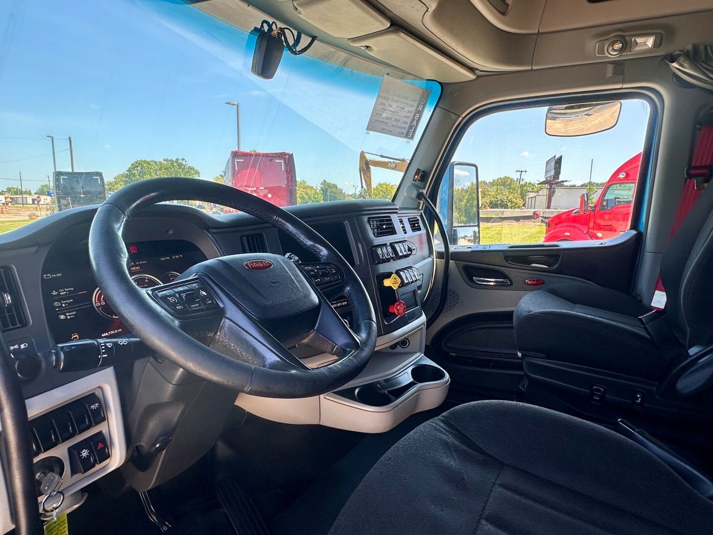 Peterbilt 579 truck interior showing steering wheel, dashboard, and driver's seat with center console; windshield view of exterior trucks.