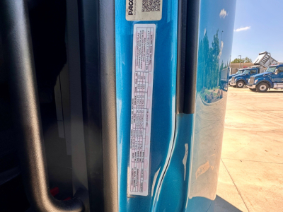 Close-up view of a blue Peterbilt truck door jamb with a vertical sticker containing vehicle information labels, text includes VIN barcodes and small print; portion of the cab and exterior reflect in the background.