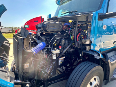 Peterbilt 579 engine bay view showing diesel engine with hoses, belts, and wiring harness; blue cab, front radiator area visible.
