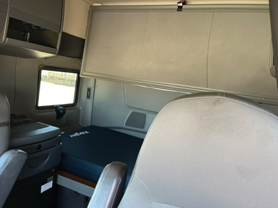 Truck cab interior showing sleeper berth area with overhead storage and window; captain seat in foreground; storage compartments mounted along wall.