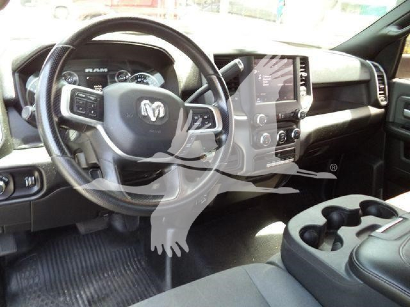 Ram truck interior showing steering wheel with RAM emblem, center touchscreen, dashboard controls, and cup holders in foreground.