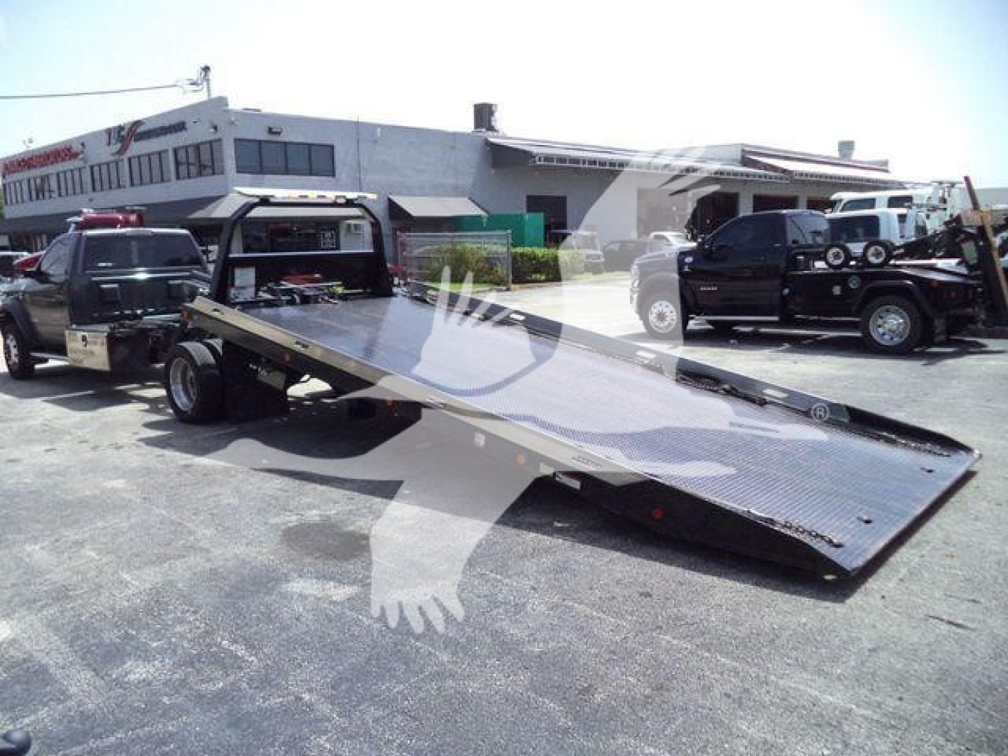 Ram 5500 rollback tow truck with a tilted flatbed in a dealership lot; body black, surrounding vehicles and building in background.