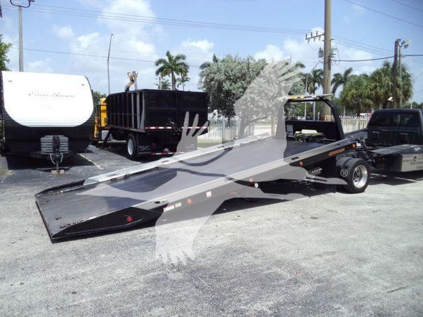 Ram 5500 rollback tow bed flatbed with hook and ramps, black metal surface, urban lot with other trucks and equipment in background.