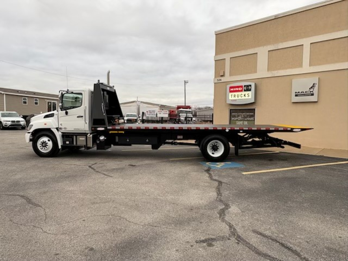Truck bed with flatbed tow truck in-service profile at dealer lot, 2025 Hino L7. Side view showing long flatbed and cab, dealership building in background.