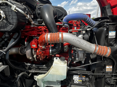 Peterbilt engine bay with red engine block, large intake hose, silver exhaust pipe with orange bands, coolant reservoir, and various hoses and components visible.