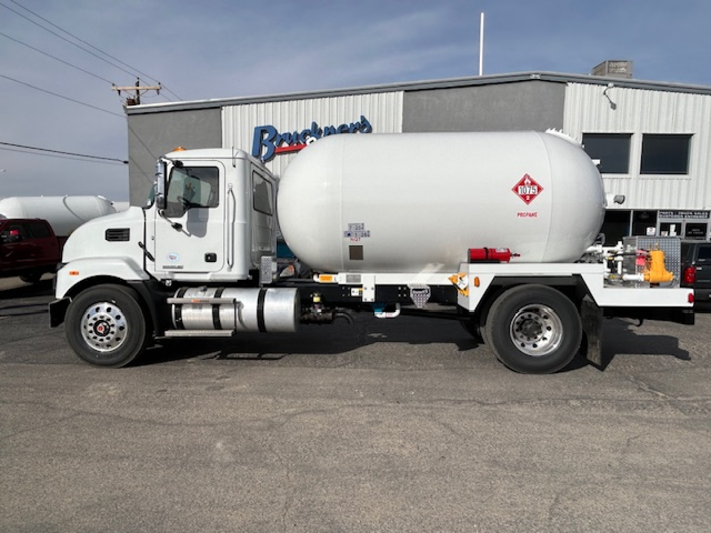 Truck-mounted liquid propane tank with Mack MD7 cab, side view, white cylindrical tank on chassis, dealership building in background.