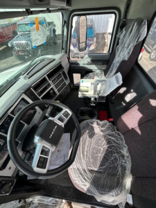 Truck cockpit interior showing steering wheel, driver's seat wrapped in plastic, dashboard with instrument cluster, and auxiliary controls in cabin.