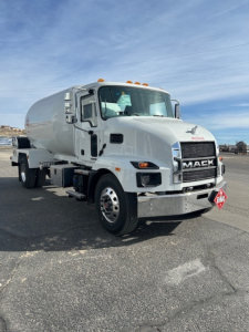Mack MD7 tanker truck in a parking lot, front three-quarter view, white cab with chrome bumper, orange marker lights, and a diamond placard on the front.