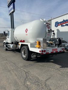 Tank truck rear view with cylindrical white tank, hazard diamond placards, rear bumper, and hoses. Visible markings include a red hazard diamond on tank, triangular warning, and equipment mounted on back.