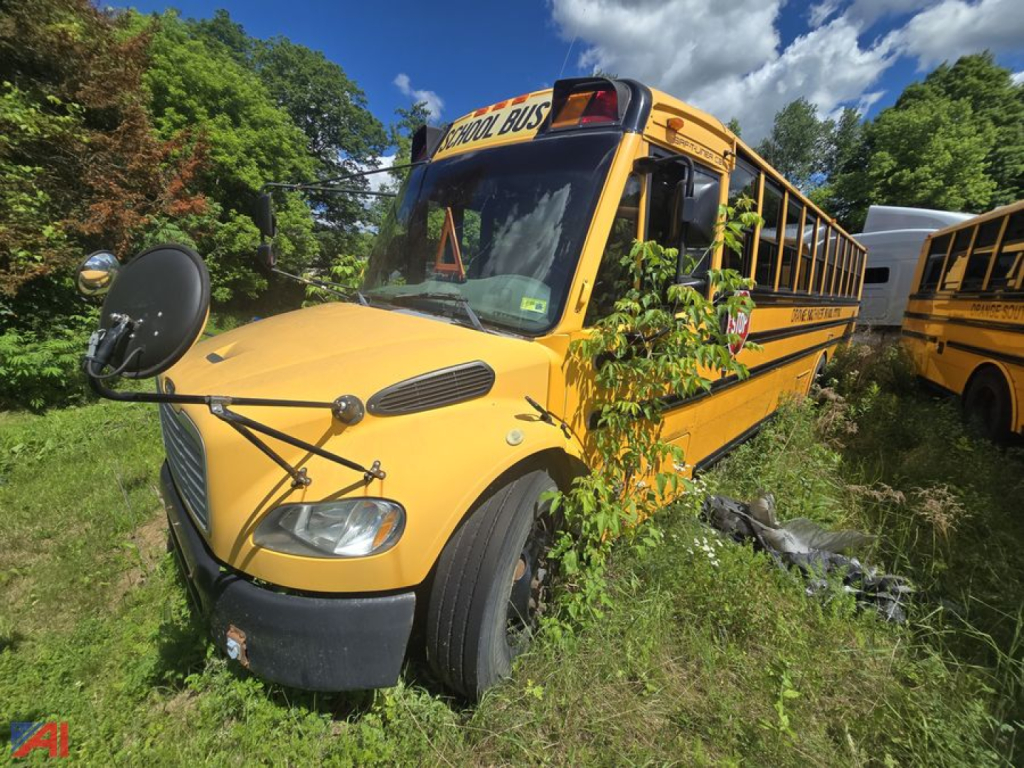 Bus front view of a yellow 2014 Freightliner B2 school bus with large mirrors, black bumper, and overgrown grass around it.