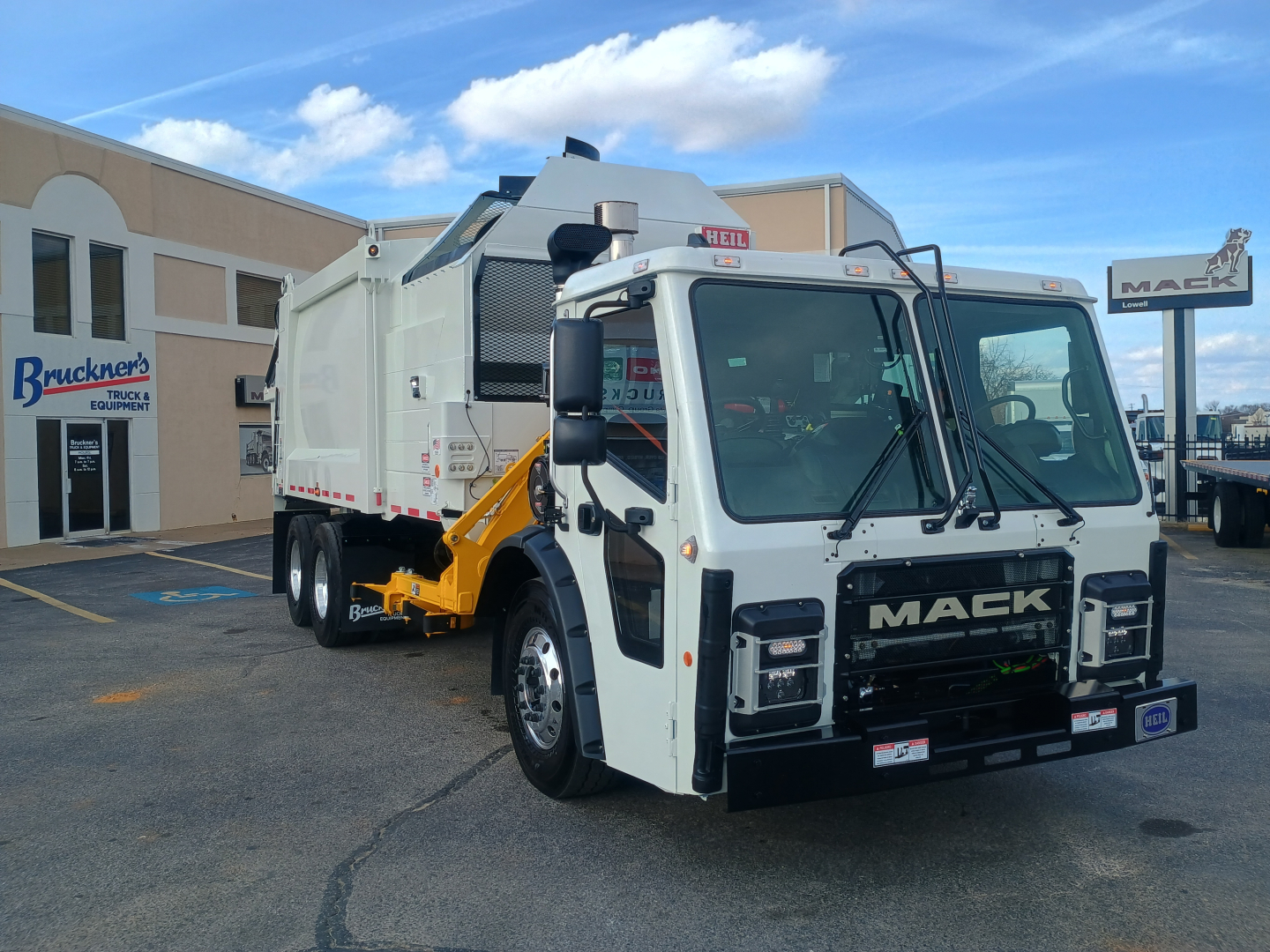 Mack LR64 refuse truck in dealership lot, front cab view with white exterior, black grille, yellow arm, and service body on rear; VIN plate not visible.