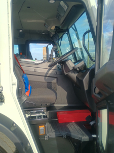 Truck cab interior of a 2026 Mack LR64 side loader showing steering wheel, driver seat, dashboard, pedals, and red toolbox under dash.