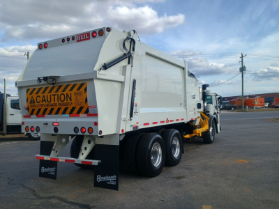 Mack LR64 refuse truck rear-side view showing rear dump body with caution sign, dual rear axles, and side loader truck chassis.