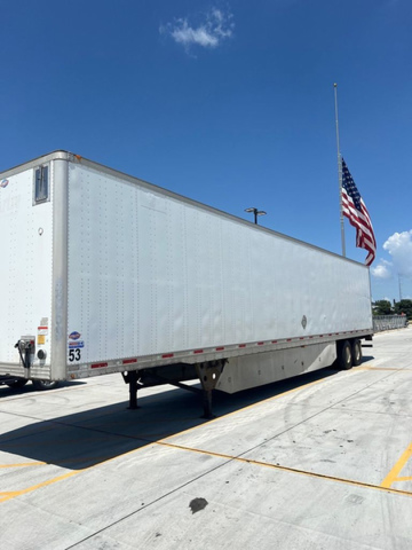 Long white utility van trailer in a parking lot with a flag and blue sky, rear and side views visible.