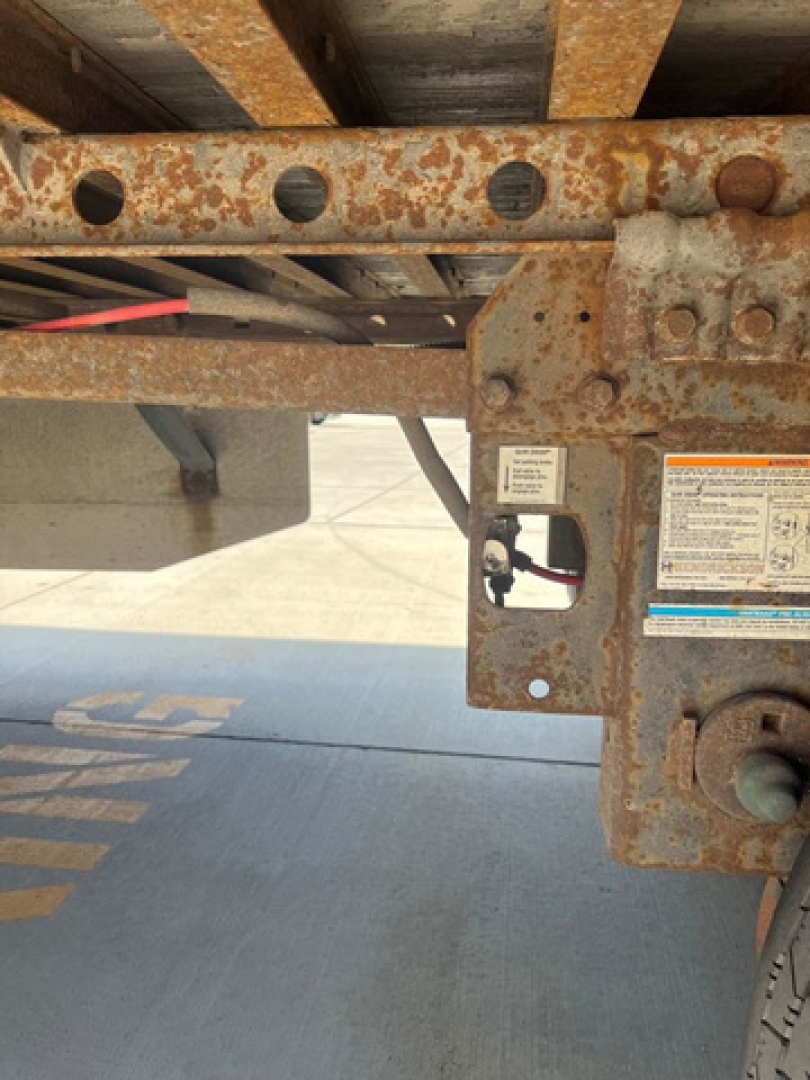 Rusty trailer frame with orange corrosion and a metal bracket; visible label plate and wiring near the hitch area.