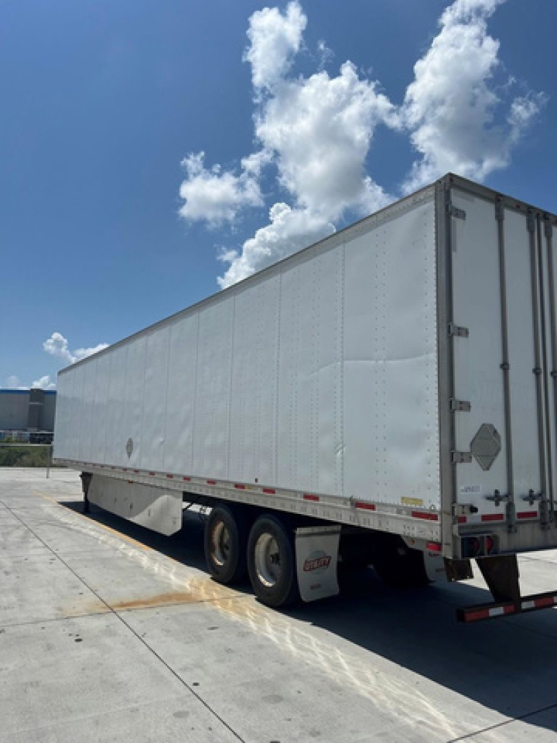 Utility van trailer showing white enclosed dry van with tandem axles and rear doors, side view, under clear sky.