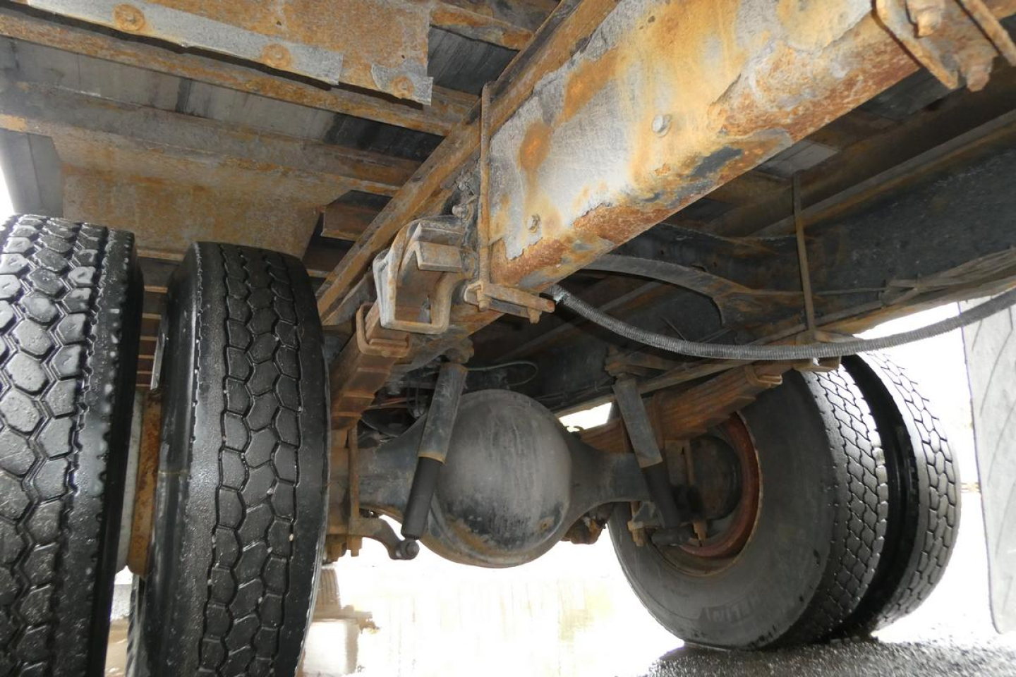 Truck rear undercarriage showing drivetrain—diff housing, dual rear axles, leaf springs, and riveted frame rails with rust-colored residue.