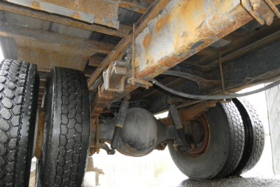 Truck rear undercarriage showing drivetrain—diff housing, dual rear axles, leaf springs, and riveted frame rails with rust-colored residue.