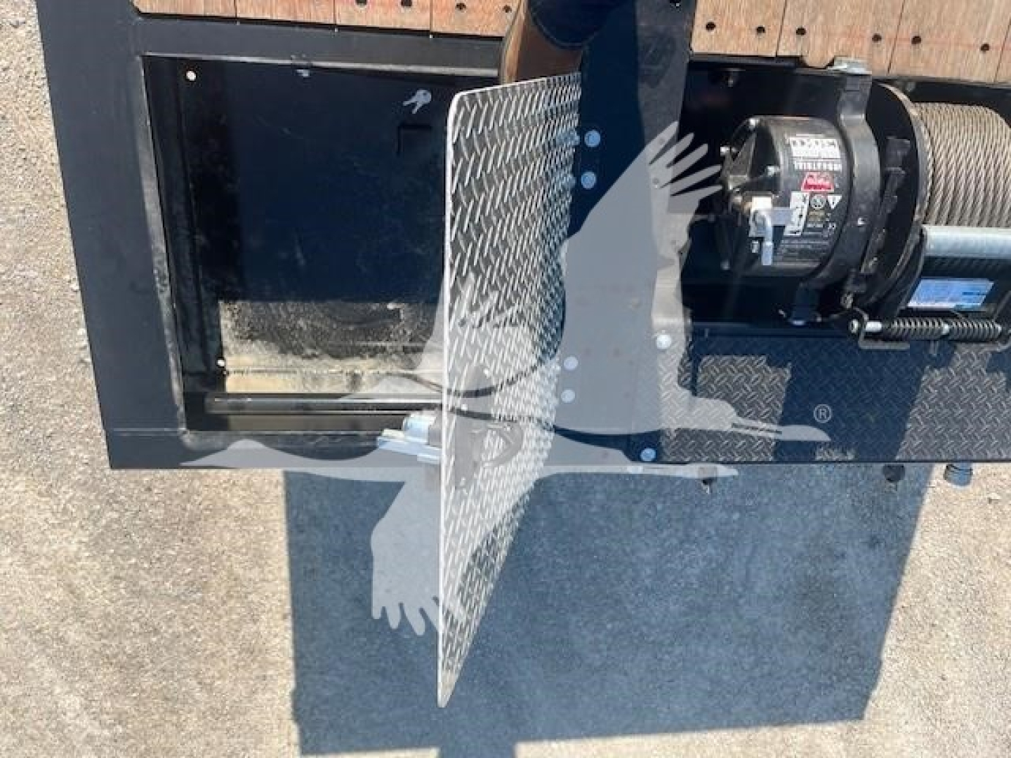 Trailer component with a winch motor and a metal diamond-plate guard, showing part of a trailer frame and equipment.