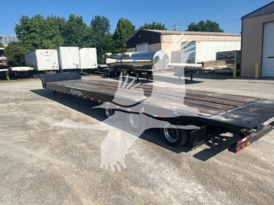 Low-angle view of a flatbed trailer with a long steel deck, dual rear axles, and a visible hitch, in an outdoor lot with other trailers and a warehouse in the background.