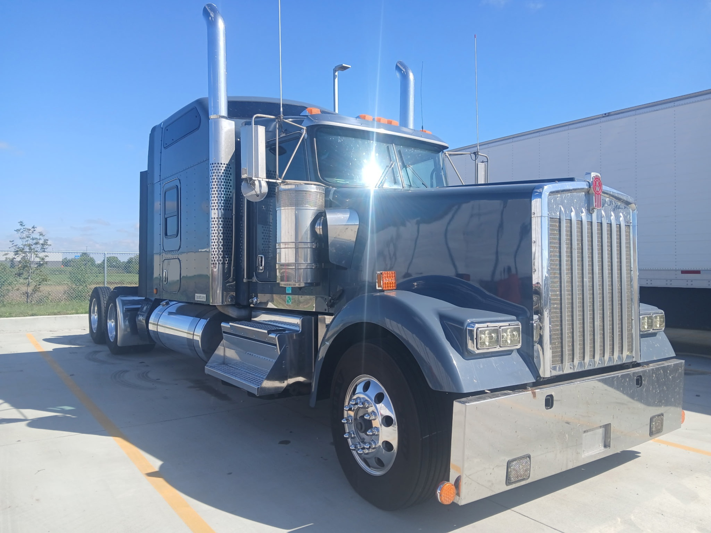 Kenworth W900L conventional sleeper truck front-left view showing chrome grille, dual exhaust stacks, large chrome bumper, and fuel tanks.