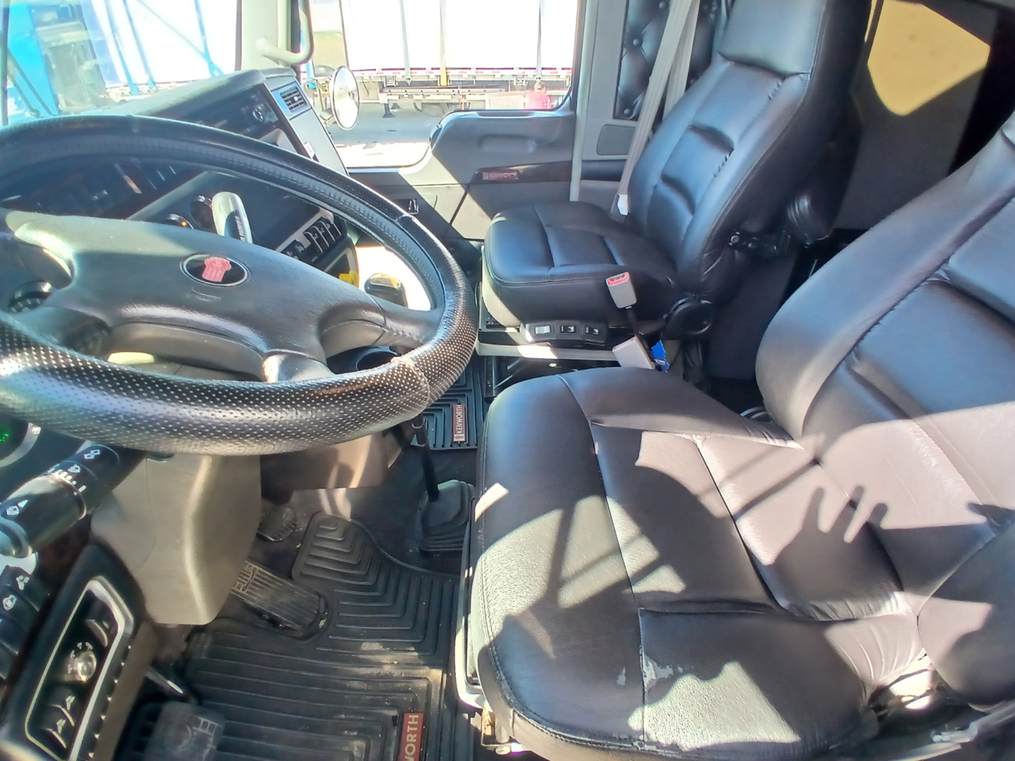 Kenworth W900L truck interior showing steering wheel, driver seat, and front passenger seat with dash area and floor mats visible.