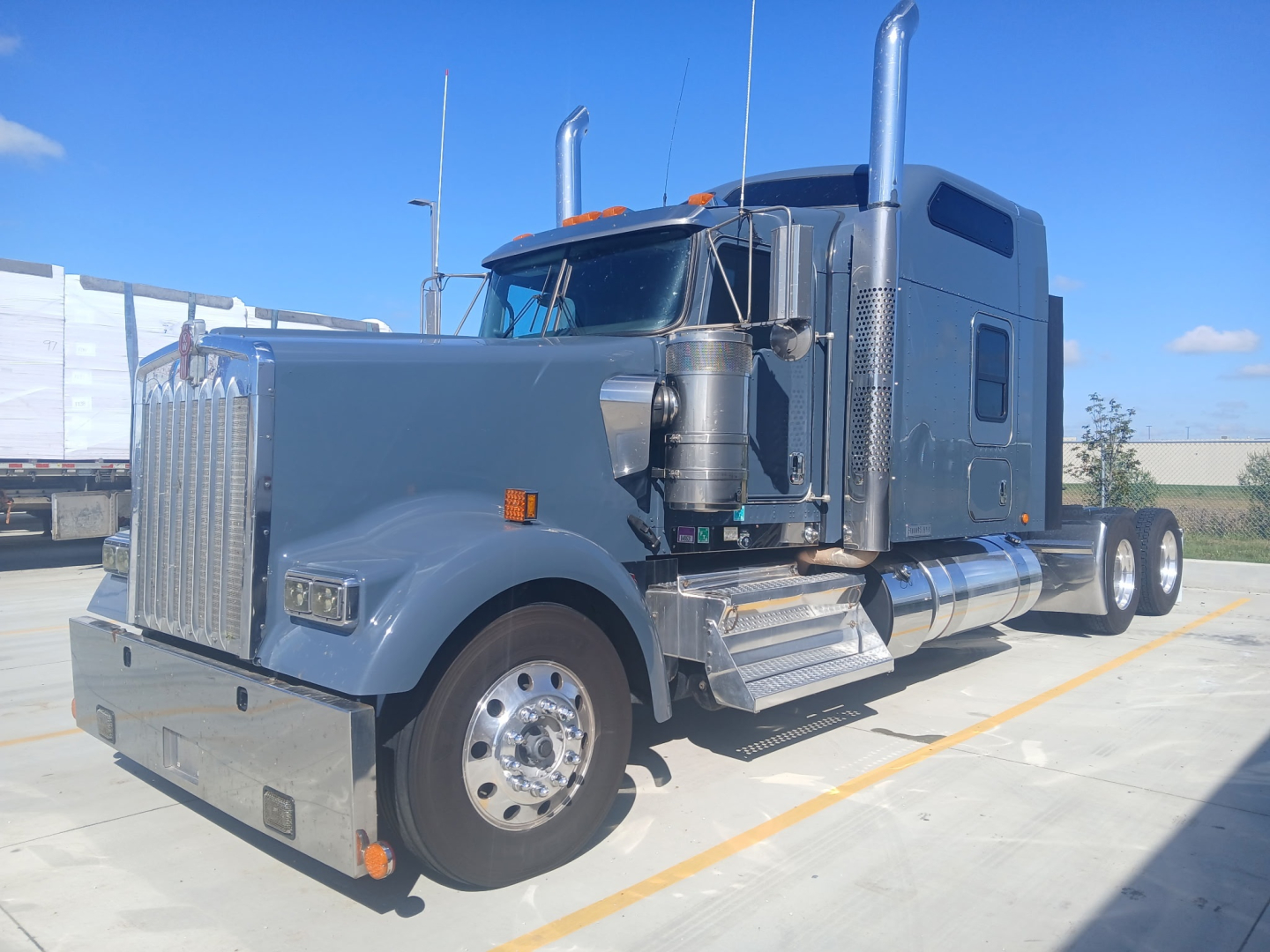 Kenworth W900L conventional sleeper truck in blue-gray paint, front-left view, with large chrome grille, dual exhaust stacks, and polished fuel tanks.
