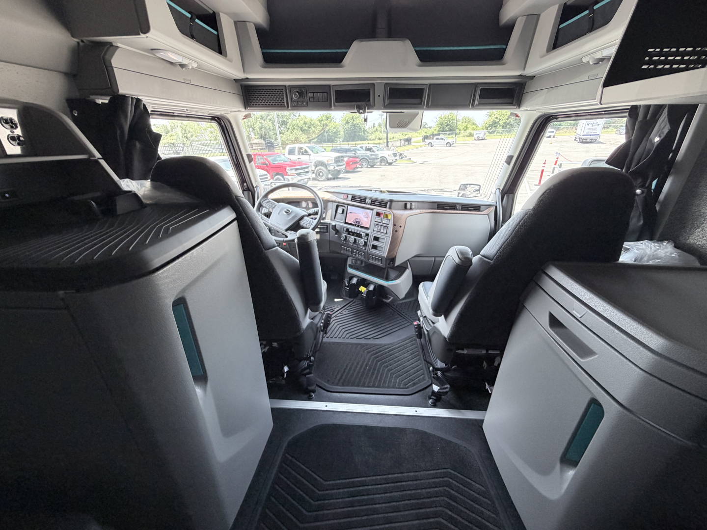 Truck cab interior showing steering wheel, driver and passenger seats, dashboard with gauges and controls, overhead compartments, and steps leading into the sleeper area.