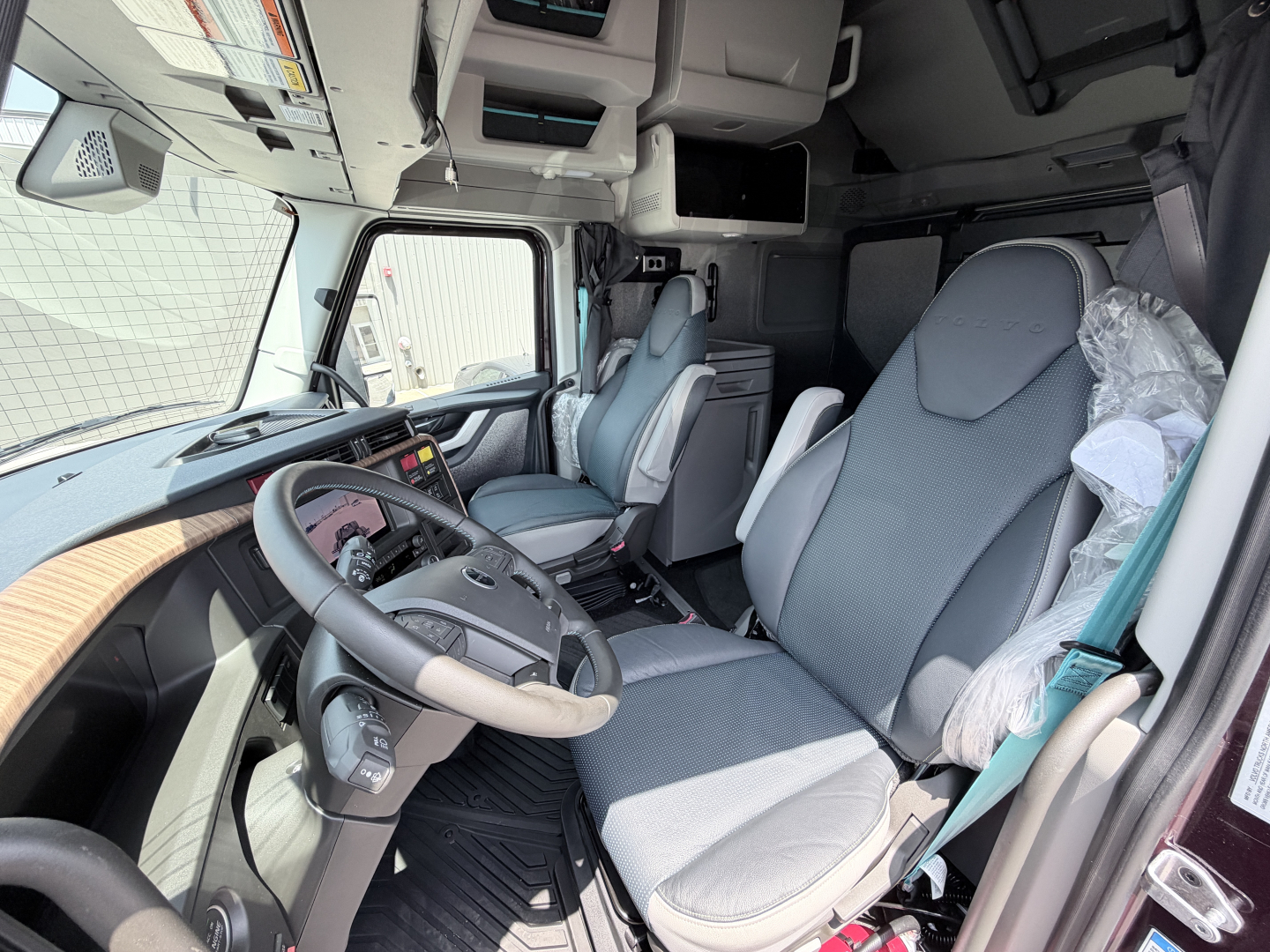 Truck cab interior in view showing driver seat and dashboard with large steering wheel and overhead compartments.