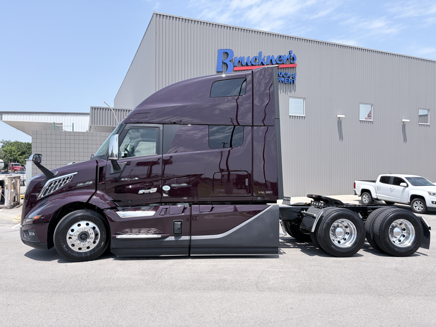 2027 Volvo VNL64T860 tractor-trailer in maroon color, side view, with sleeper cab and dual rear axles, parked in front of a building with Bruckner sign.