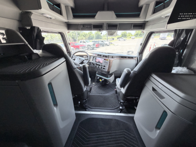 Truck cab interior showing steering wheel, driver and passenger seats, dashboard with gauges and controls, overhead compartments, and steps leading into the sleeper area.