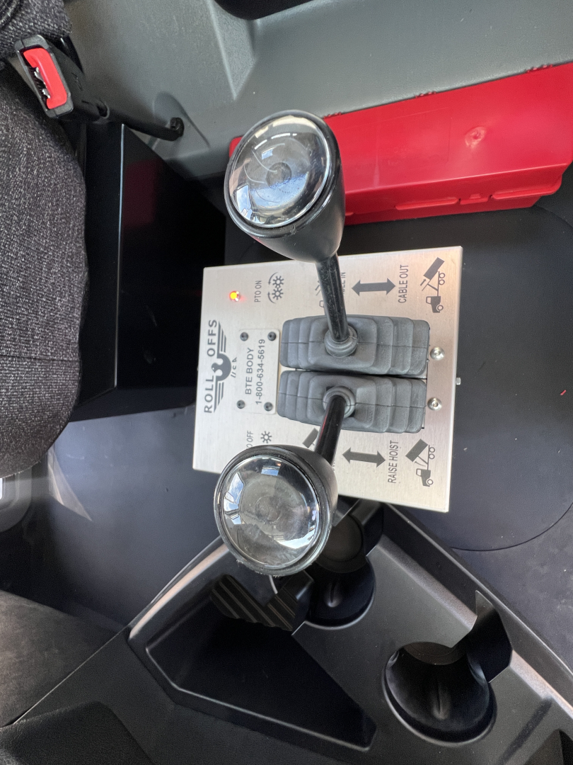 Mack GR64B gear shift console showing two control levers with round knobs and labeled switch panel. Visible text includes 'ROLL OFFS' and symbols on the control plate.