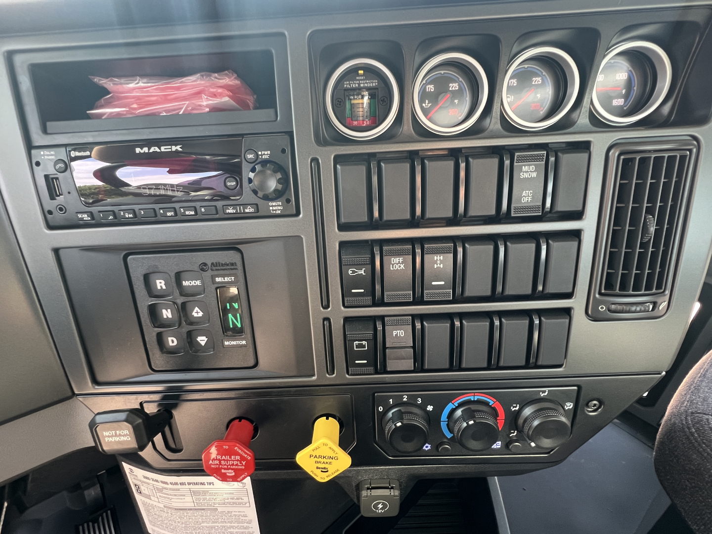 Dashboard of a Mack truck cab showing steering column, gauges, switches, and radio; red and yellow caution tags hang from panel; climate controls and ignition area visible.