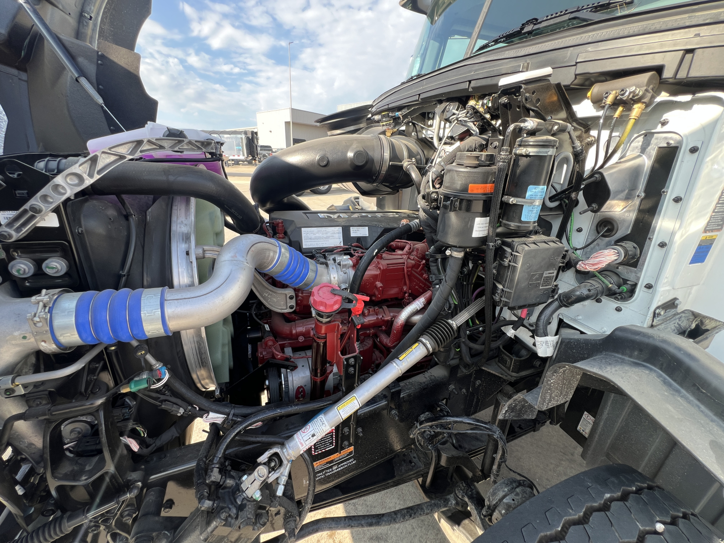 Mack GR64B engine bay showing red engine block, various hoses, wiring, and components in a truck front compartment.