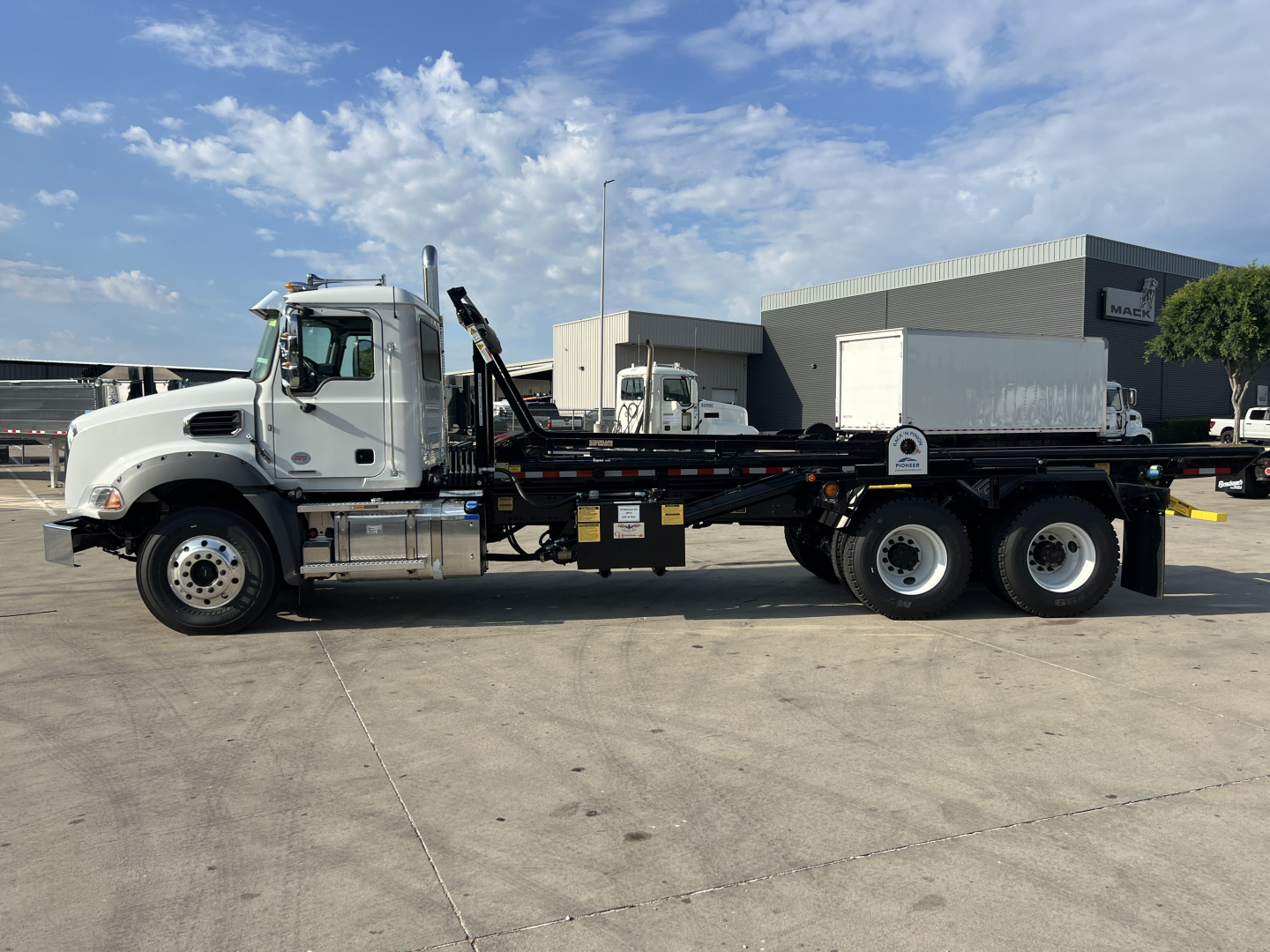Side view of a 2026 Mack GR64B roll-off truck showing a white cab and long black flatbed with dual rear axles.