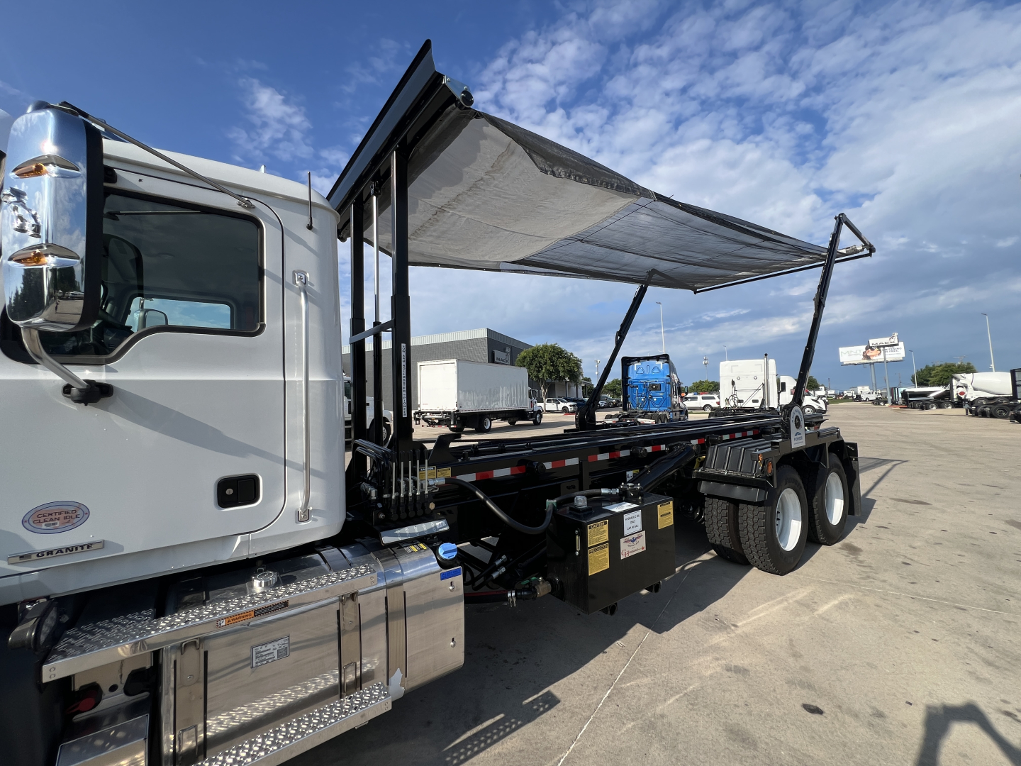 Mack GR64B roll-off truck side view showing side cab, long chassis, dual rear axles, and elevated tarp/haul bed. Visible are chrome fuel tanks, steps, and black tool/utility boxes along the frame.