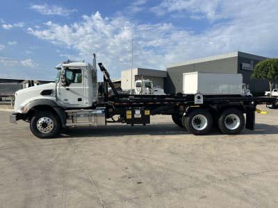 Side view of a 2026 Mack GR64B roll-off truck showing a white cab and long black flatbed with dual rear axles.
