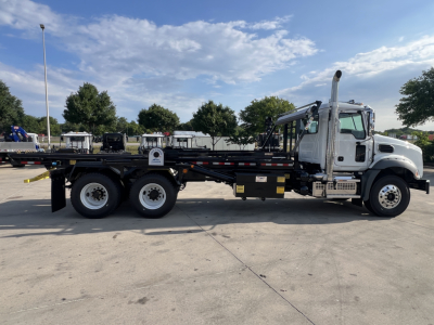 Mack GR64B rolloff truck shown in profile: white cab, long flatbed with side rails, dual rear axle, chrome exhaust stack, and heavy-duty chassis.