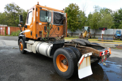 Orange Freightliner Cascadia 125 daycab tractor showing side and rear chassis with fuel tank and hitch. Visible components: cab, fuel tank, exhaust, rear wheels, mud flaps, and hydraulic lines.