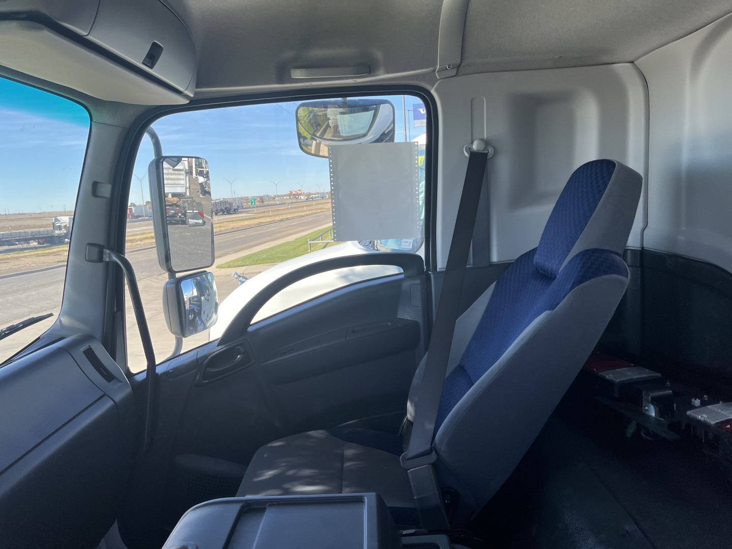 Isuzu FTR truck interior cabin front seats and dashboard visible with blue-and-gray seat upholstery, steering wheel, side window mirror, and driver seat belt.
