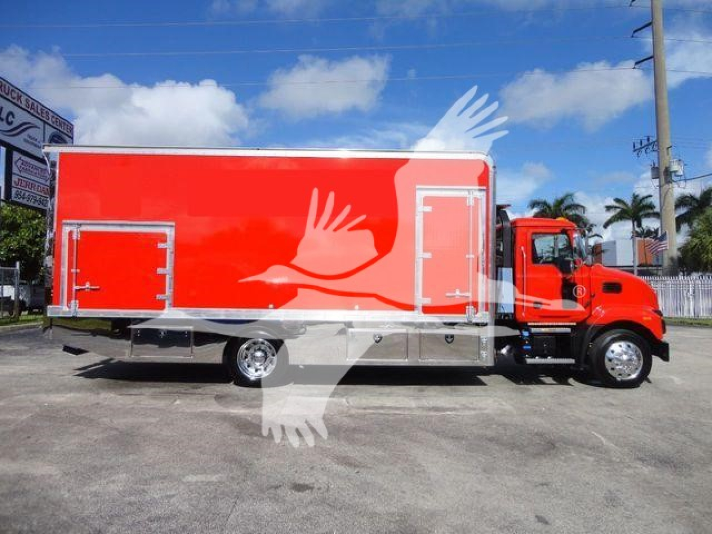 A bright red Mack MD6 rollback truck with a long cargo box, showing white graphic door panels and a white bird graphic overlay on the side.
