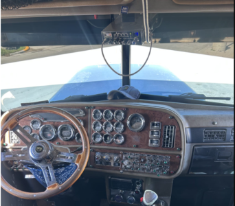 Truck cab interior showing steering wheel and dashboard gauges in a Peterbilt 379 EXT, wood grain dash, numerous analog gauges, overhead electronics, and a windshield view.