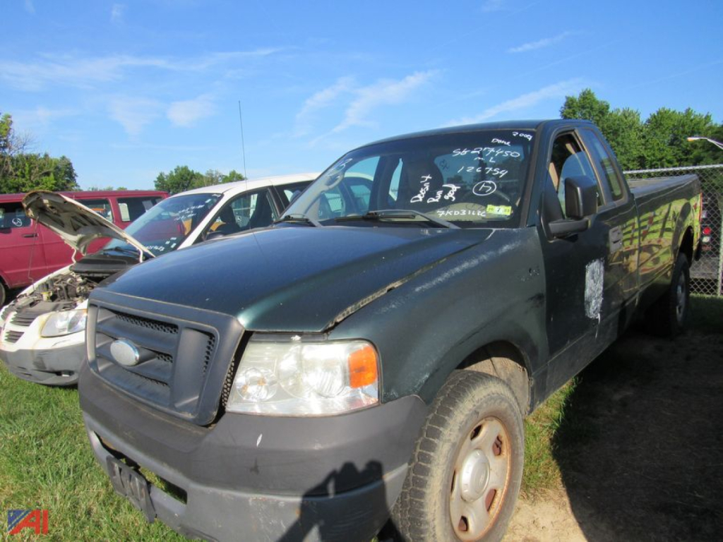 Ford F-150 pickup truck 2007 model in a used lot, dark green body, front-left view, writing on windshield, cargo bed, other vehicles in background.